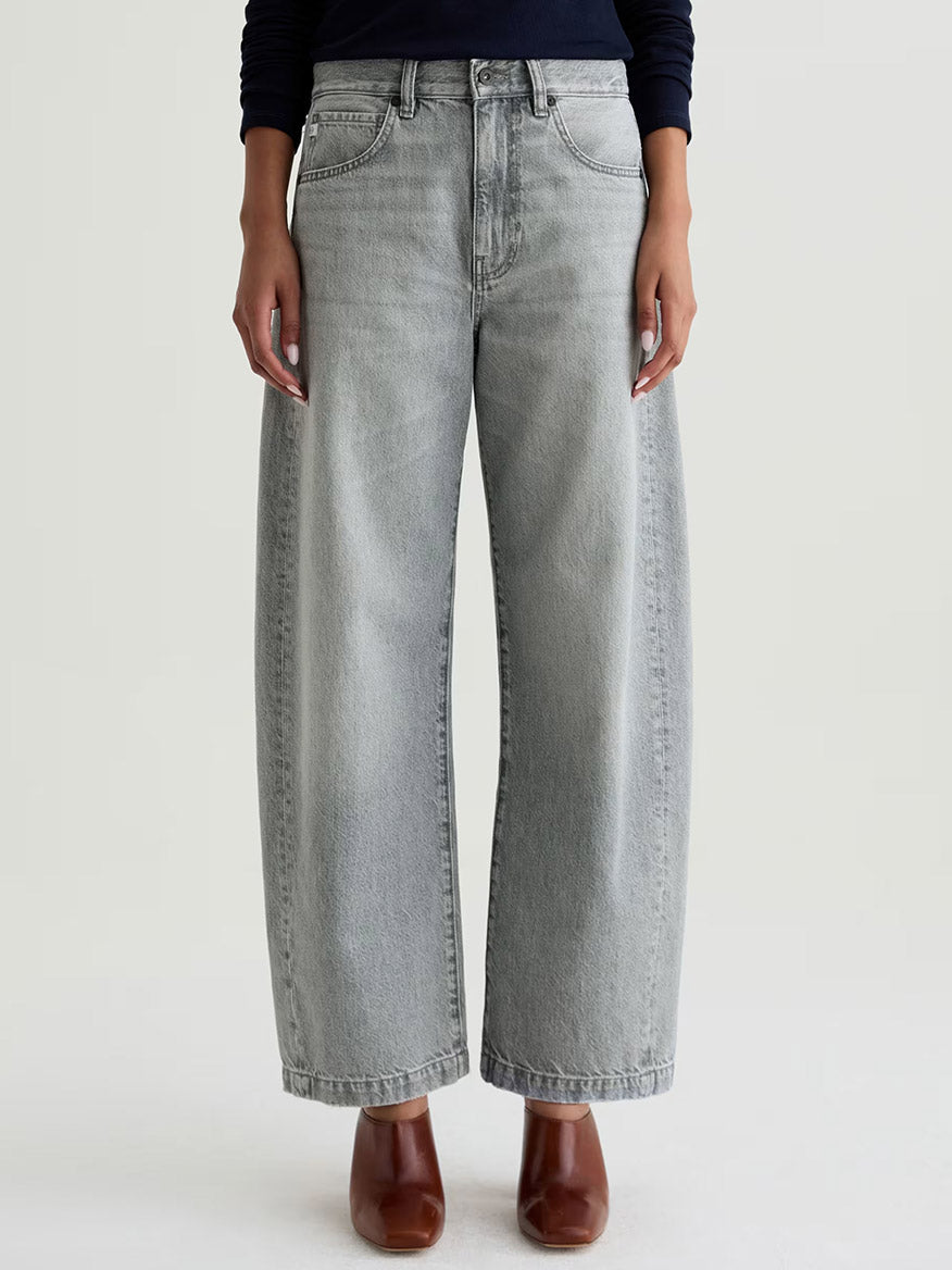 Wearing AG Jeans Hattie High Rise Barrel in Al Fresco—petite-friendly, light gray, high-waisted wide-leg non-stretch denim—with brown heeled shoes, standing against a plain background.