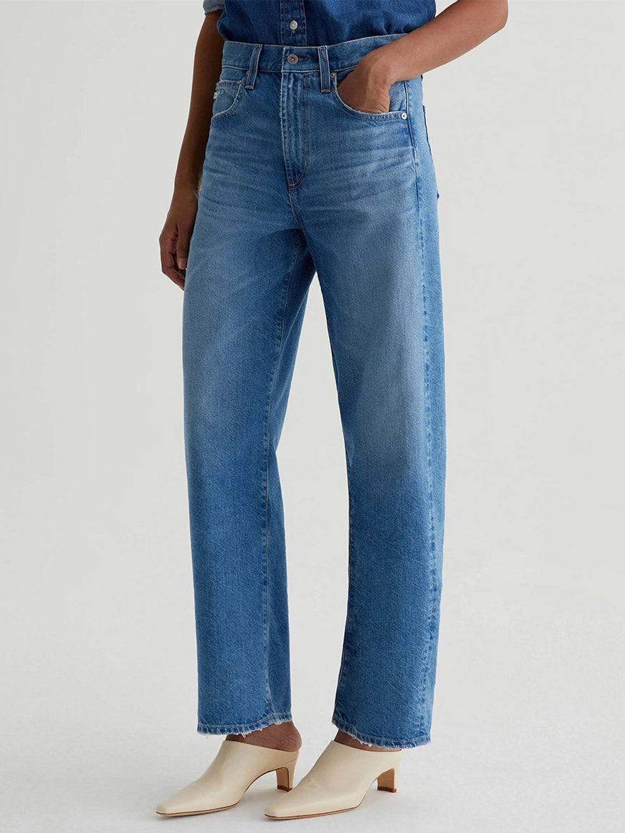 Model wearing AG Jeans Hattie High Rise Barrel in Flashback, styled with a tucked-in denim shirt and cream heeled shoes, standing against a plain light background.