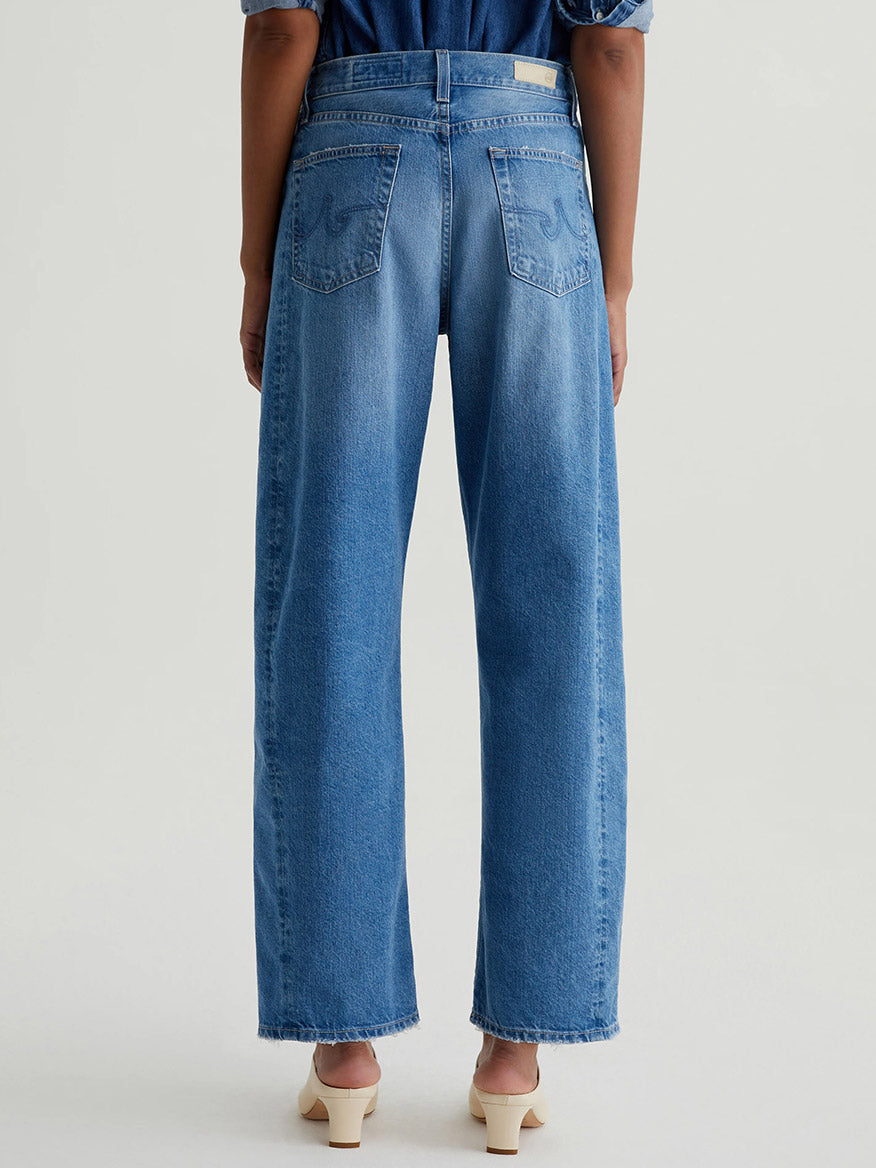 Back view of a person wearing AG Jeans Hattie High Rise Barrel in Flashback, a brilliant blue shade, paired with beige heeled sandals, standing against a plain light background.