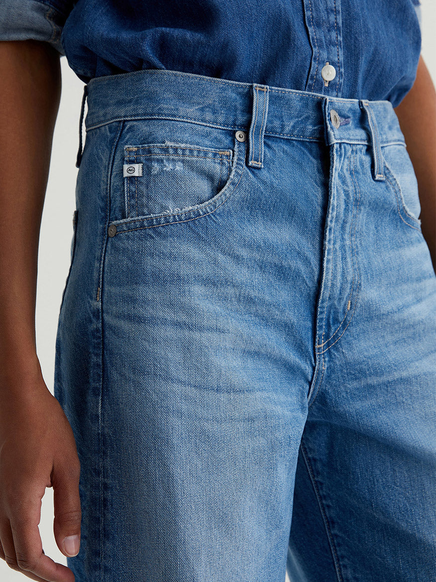 Model in AG Jeans Hattie High Rise Barrel in Flashback, paired with a matching denim shirt, shown from waist to mid-thigh with one arm at the side.
