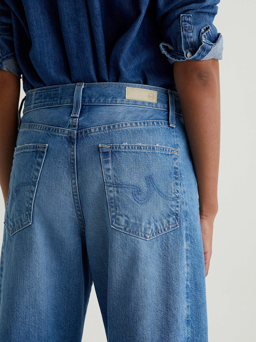 Model shown from the back wearing AG Jeans Hattie High Rise Barrel in Flashback, paired with a denim shirt with rolled sleeves.