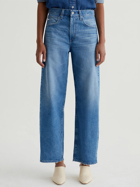 A person stands against a plain light background wearing AG Jeans Hattie High Rise Barrel in Flashback, paired with a tucked-in blue shirt and cream shoes.