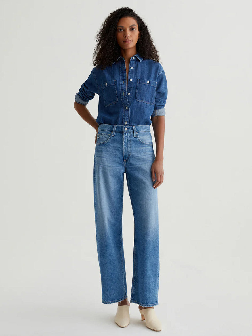 A woman faces forward, wearing the AG Jeans Hattie High Rise Barrel in Flashback with a denim shirt tucked in and cream shoes.
