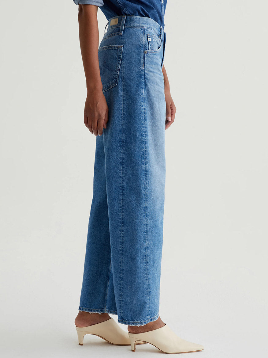 A person stands sideways against a plain background wearing AG Jeans Hattie High Rise Barrel in Flashback, paired with a blue shirt and off-white heeled mules.