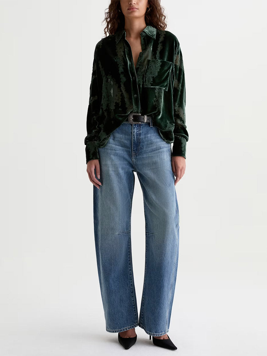 A person stands against a plain background wearing a loose dark green velvet shirt, the AG Jeans Mercer Cuffed High-Rise Barrel Jean in Vibrato, a black belt, and black pointed-toe shoes.