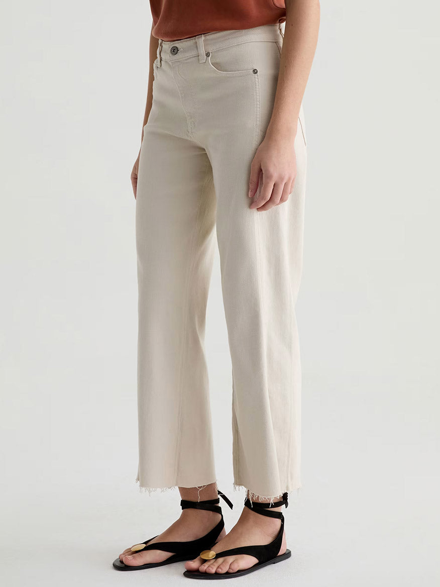 Wearing AG Jeans Saige Wide Leg Crop With Panel in Opal Stone, a brown top, and black sandals with gold accents, this person stands against a plain background.