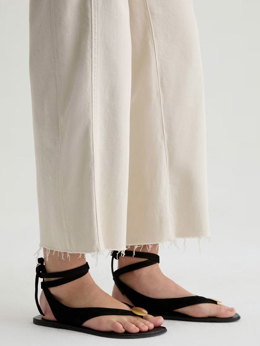 A person wears the AG Jeans Saige Wide Leg Crop With Panel in Opal Stone—cream-colored, raw stretch denim pants with frayed hems—paired with black open-toe sandals with ankle straps and a gold accent, standing on a neutral background.