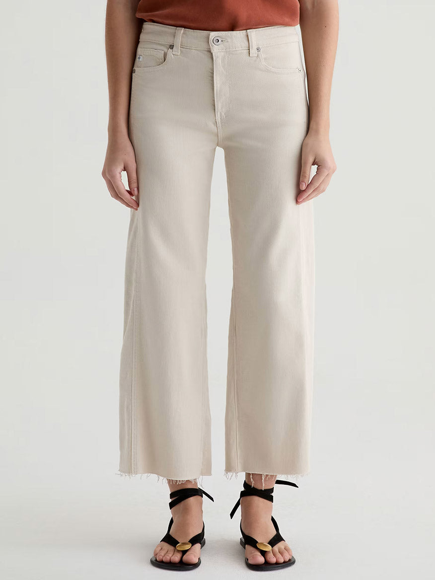 A person wearing AG Jeans Saige Wide Leg Crop With Panel in Opal Stone, crafted from raw stretch denim with raw hems, paired with black strappy sandals and standing against a light background.