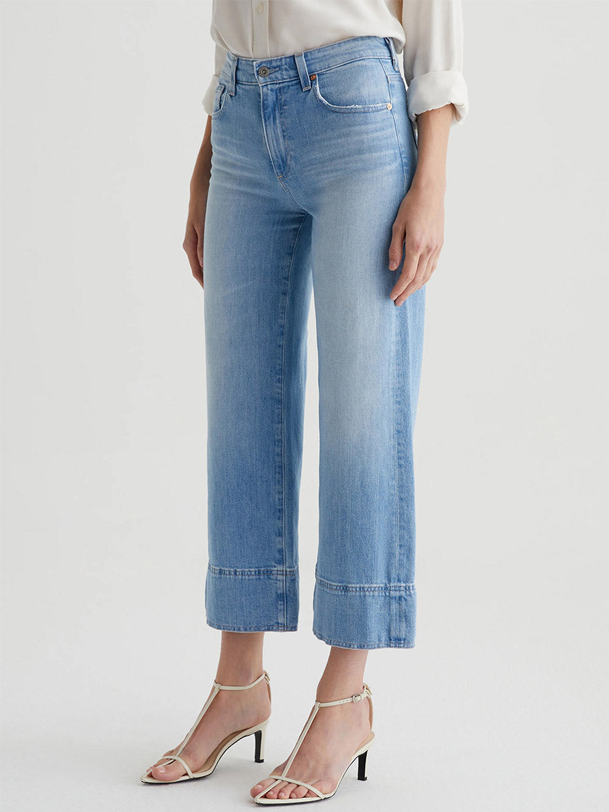 A person wearing AG Jeans Saige Wide Leg Crop in Dana Point, a white blouse, and beige heeled sandals, standing against a plain background.