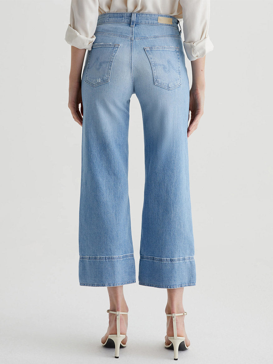 A person stands facing away, wearing AG Jeans Saige Wide Leg Crop in Dana Point, a white long-sleeve top, and white high-heeled sandals against a plain background.