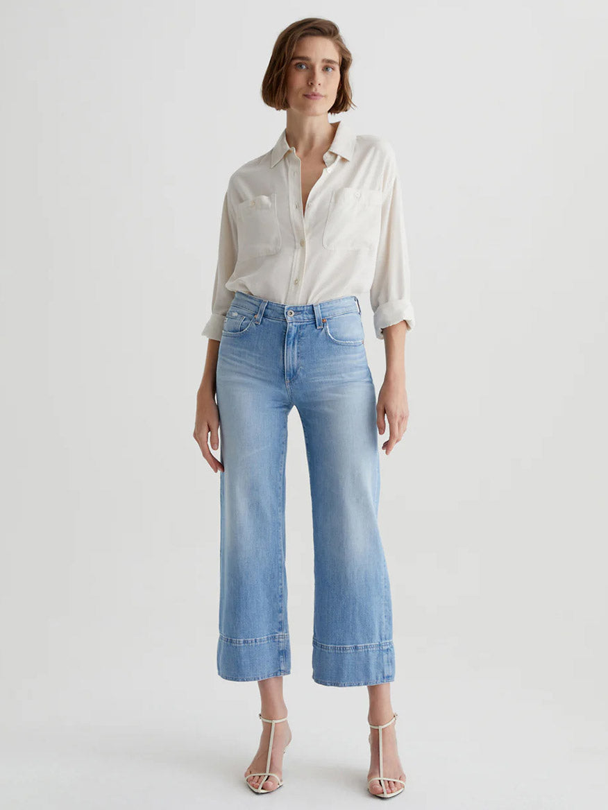 A woman stands against a plain background wearing a white button-up shirt, AG Jeans Saige Wide Leg Crop in Dana Point, and white heeled sandals.