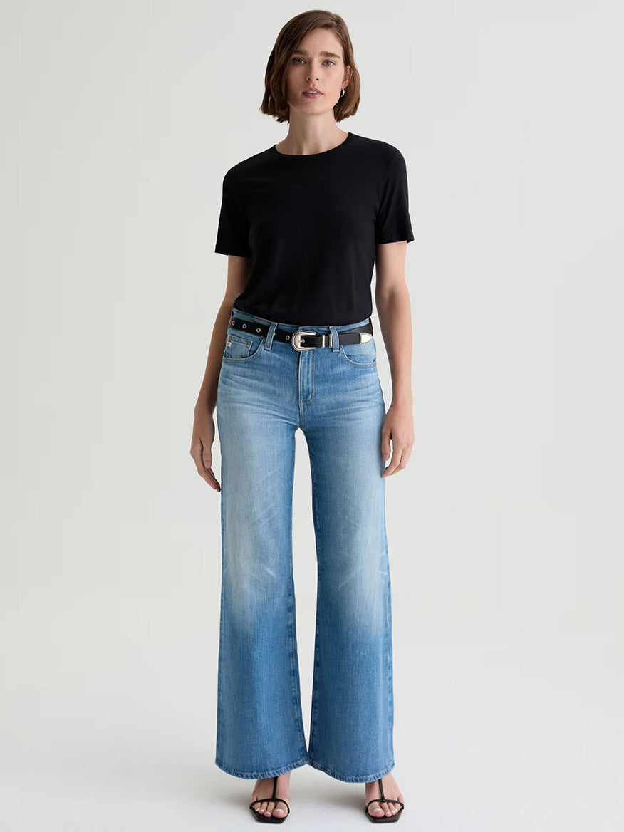 A woman faces forward, wearing a black t-shirt, AG Jeans Saige Wide Leg Mid-Rise in 18 Years Bisou premium light blue denim, a black belt, and black sandals against a plain light background.