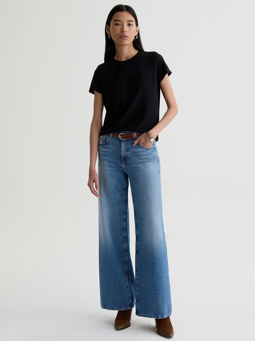 A person stands against a plain background wearing a black t-shirt, brown belt, brown shoes, and AG Jeans Saige Wide Leg Mid-Rise in American West with comfort stretch denim.