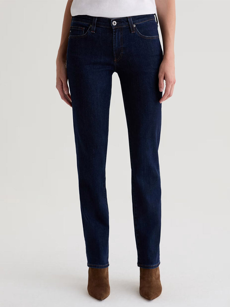 Wearing AG Jeans Sandra Low-Rise Straight in Modern Indigo, a white top, and brown suede ankle boots, a person stands against a plain light background.