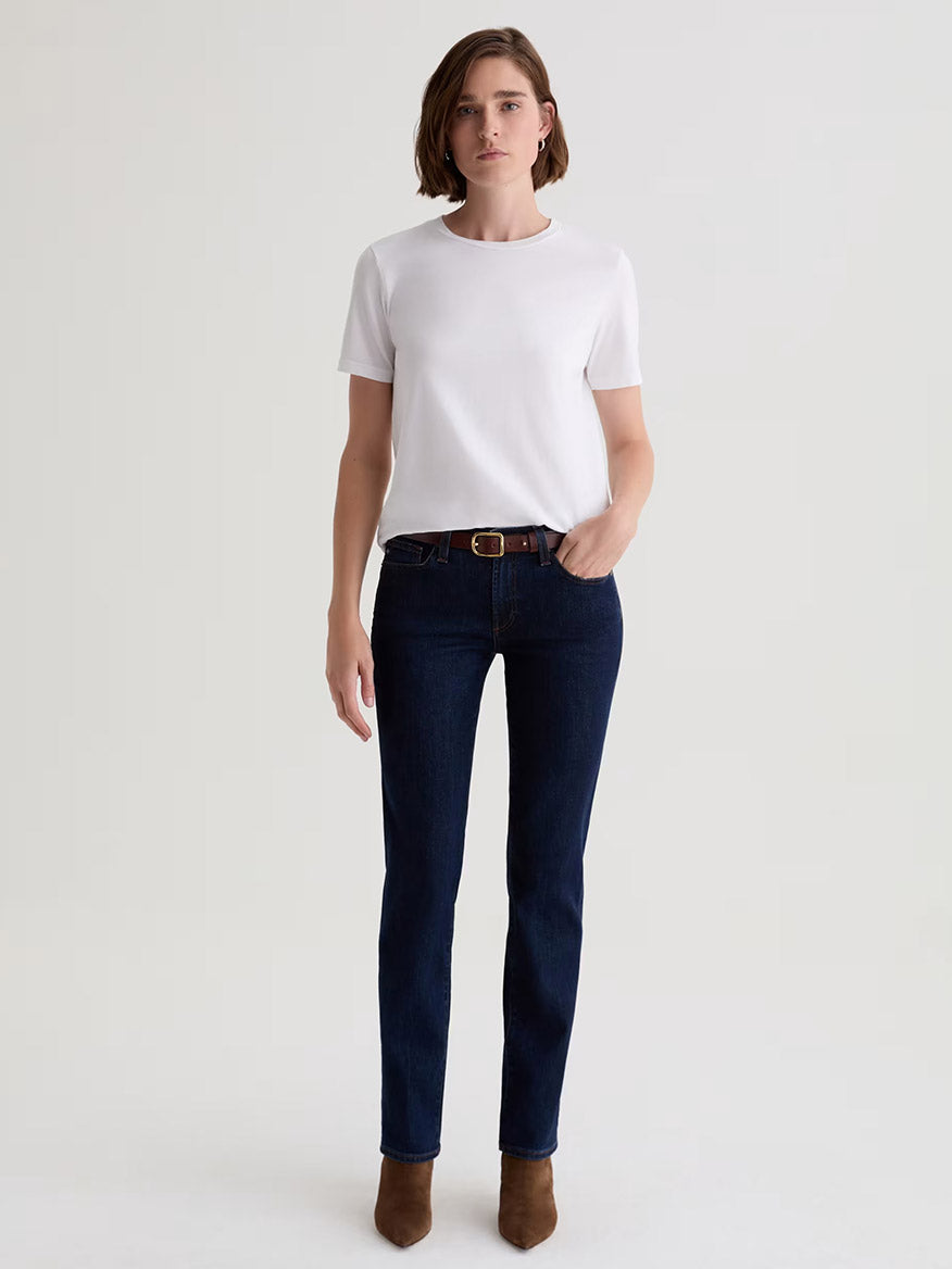 A person stands facing forward, wearing a plain white short-sleeved t-shirt, the AG Jeans Sandra Low-Rise Straight in Modern Indigo, a brown belt, and brown ankle boots against a plain light background.