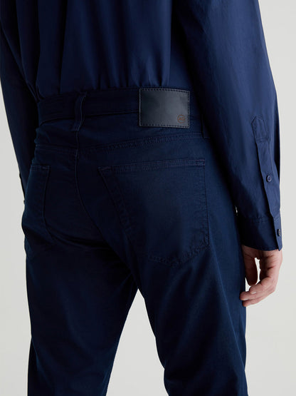 Close-up view of a person wearing AG Jeans Tellis in Bay Bridge and a matching shirt, with a focus on the back pocket stitching and brand label.