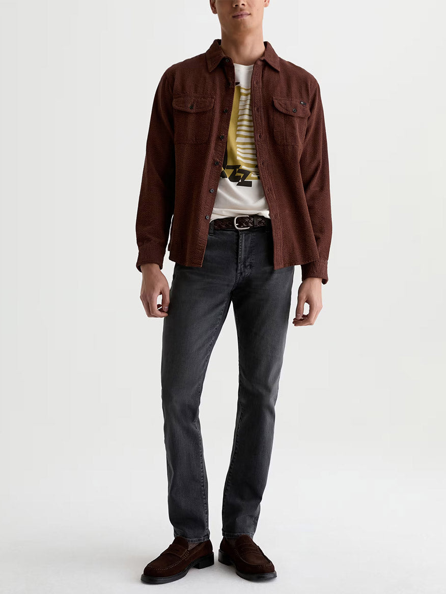A man stands against a plain background wearing a maroon button-up shirt over a graphic tee, a black belt, brown loafers, and AG Jeans Tellis in 7 Years Black Nile.