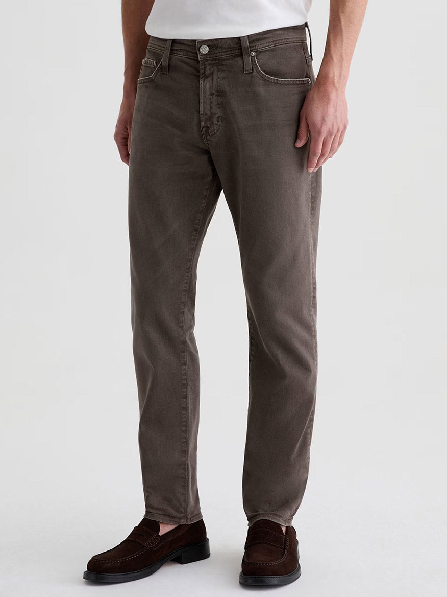 A person wearing AG Jeans Tellis in 7 Years Sulfur Brown Slate, a white shirt, and brown loafers, standing against a plain light background.