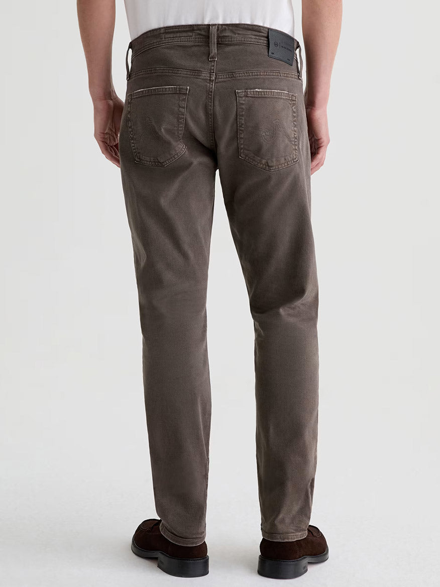 A person stands with their back to a plain light background, wearing a white shirt, dark brown shoes, and AG Jeans Tellis in 7 Years Sulfur Brown Slate.