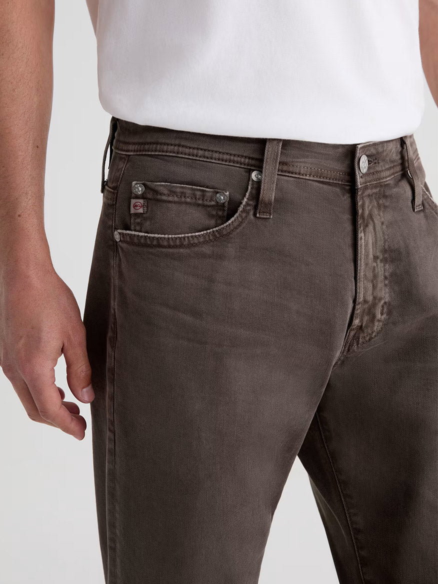 A person wearing a white shirt and AG Jeans Tellis in 7 Years Sulfur Brown Slate, shown from waist to mid-thigh.