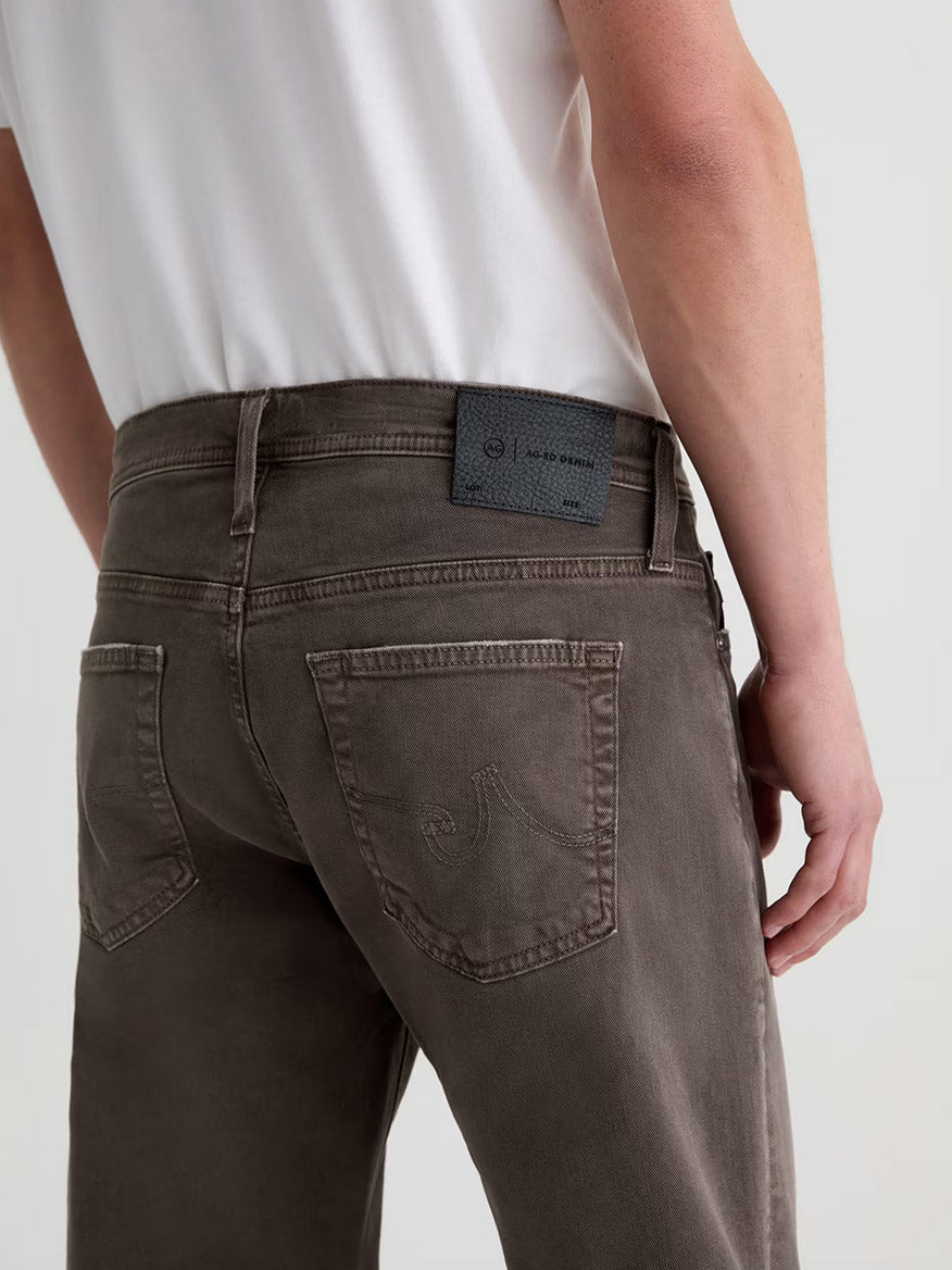 A person wearing AG Jeans Tellis in 7 Years Sulfur Brown Slate, shown from the back with focus on the rear pockets and waistband label.