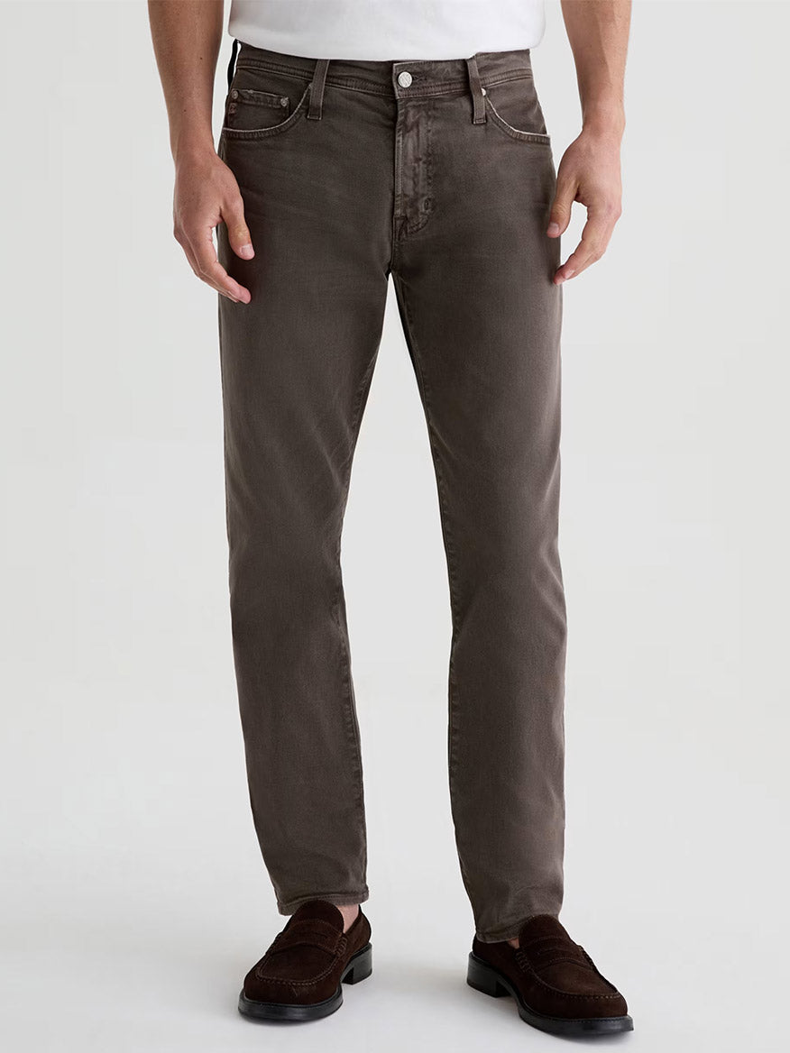 A man stands facing forward against a plain background, wearing AG Jeans Tellis in 7 Years Sulfur Brown Slate, a white shirt tucked in, and brown loafers.