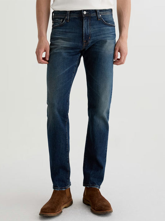 Someone wearing AG Jeans Tellis in 9 Years Alton, styled with a white shirt and brown suede boots, stands against a plain light background.