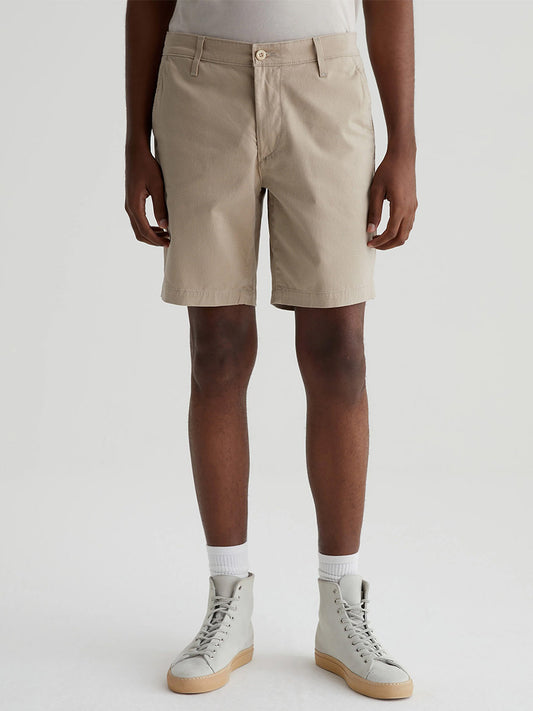 A person wearing AG Jeans Wanderer Short in Parquet Khaki Multi, white socks, and cream-colored high-top sneakers stands against a plain background.