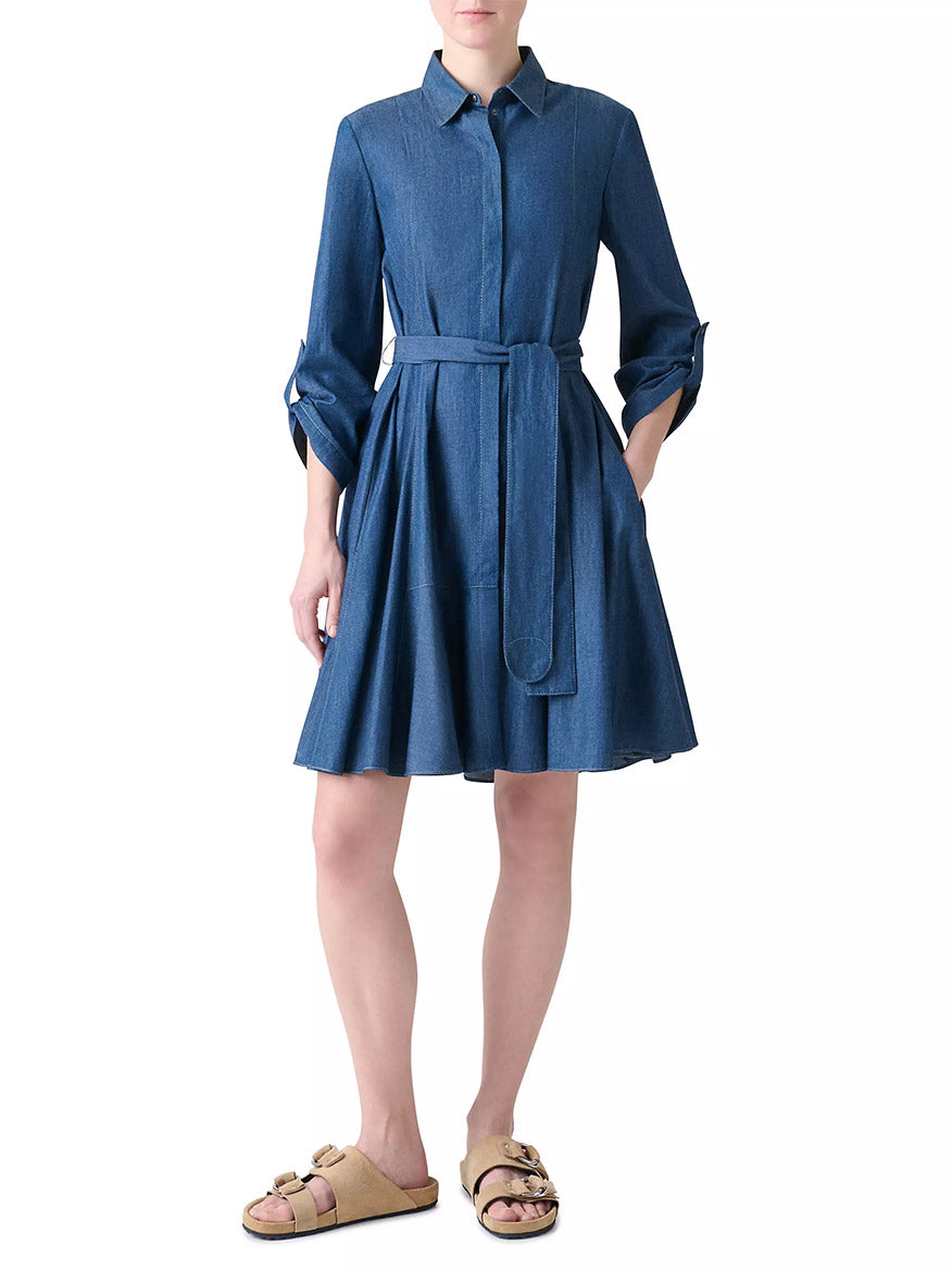 A person wearing the Akris Punto Tie-Waist Denim Shirtdress in Medium Blue with bracelet-length sleeves, a collar, and tan sandals stands against a white background.