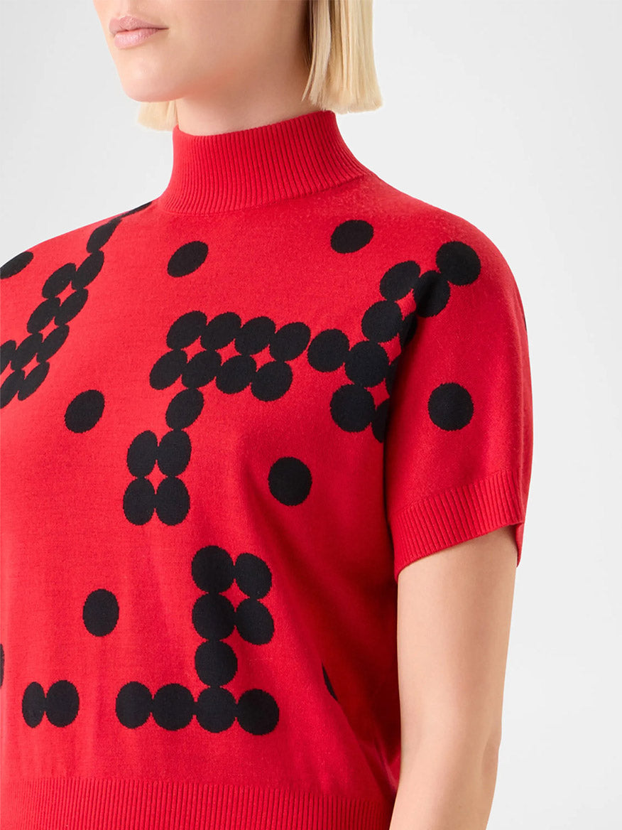 A person wears the Akris Punto Dot Grid Jacquard Knit Sweater in Vermillion/Black, featuring a red short sleeve design with black geometric dot grid pattern and a mock neckline.