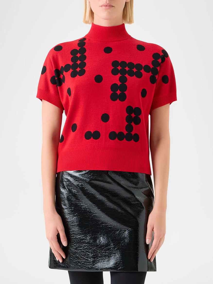 Someone stands against a plain background wearing the Akris Punto Dot Grid Jacquard Knit Sweater in Vermillion/Black, featuring black polka dot patterns, paired with a black shiny skirt.
