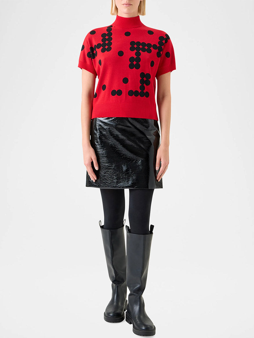 Someone wears the Akris Punto Dot Grid Jacquard Knit Sweater in Vermillion/Black with a shiny black mini skirt, black tights, and tall black boots, standing against a plain background.