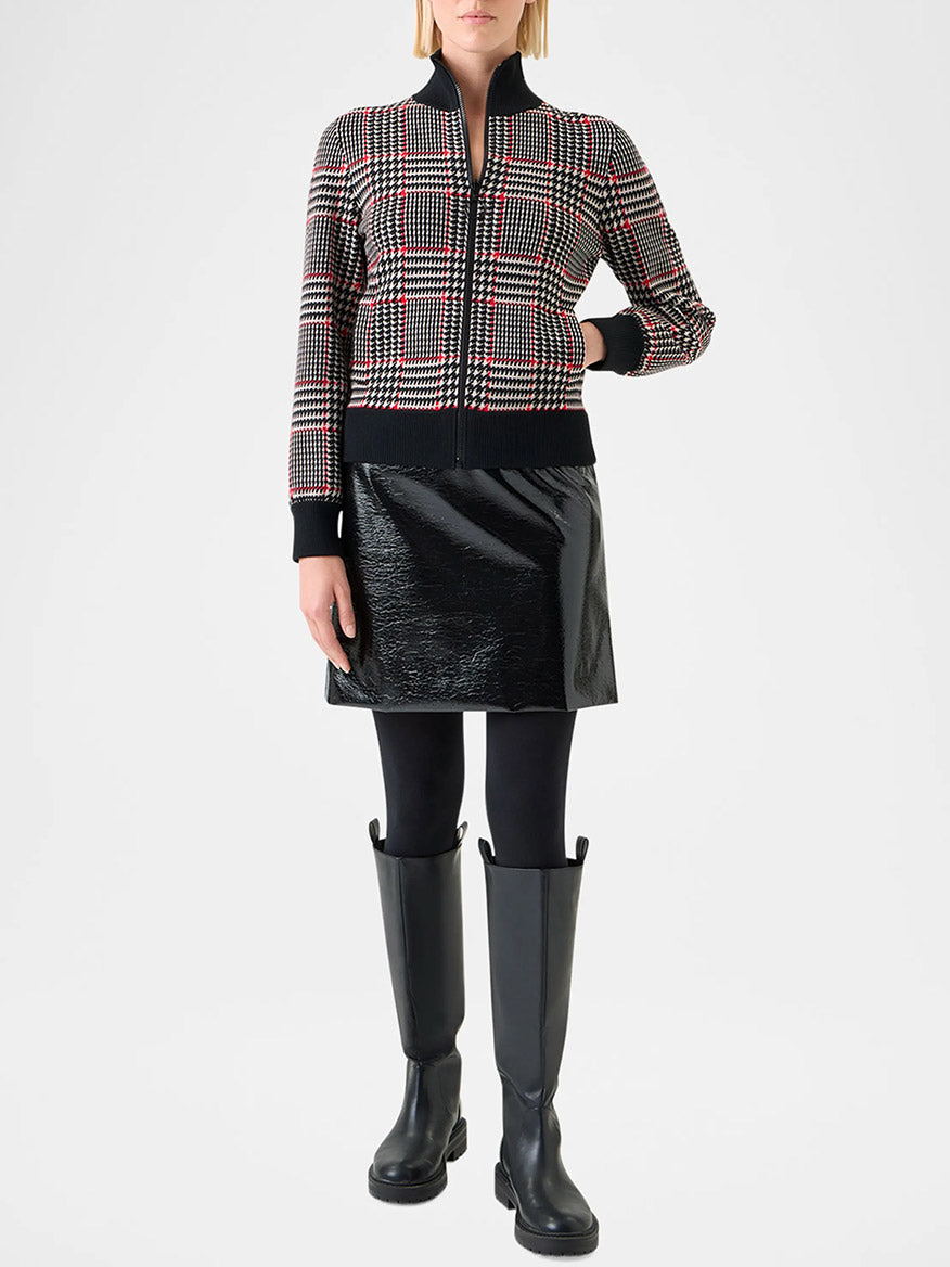 A person stands against a plain background wearing the Akris Punto Mock Neck Plaid Cardigan in Black/Cream/Vermillion, a black mini skirt, black tights, and knee-high black boots.