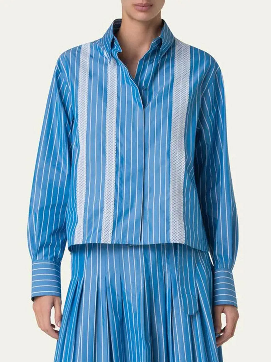 Wearing the Akris Punto Pajama Stripe Boxy Collared Shirt in Medium Blue/Cream, a person stands against a plain background paired with a matching pleated skirt.