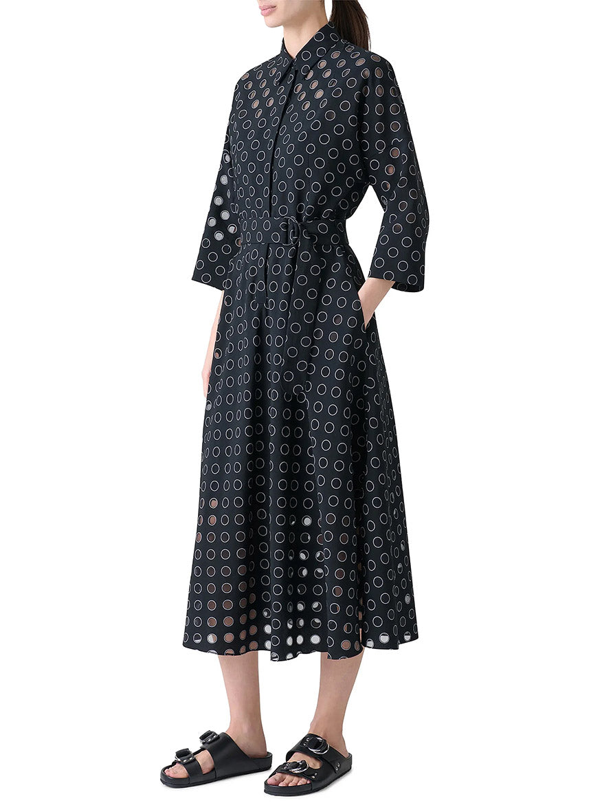 A person wearing the Akris Punto Midi Polka Dot Organzino Belted Dress in Black/Cream with black sandals, standing with hands in pockets.