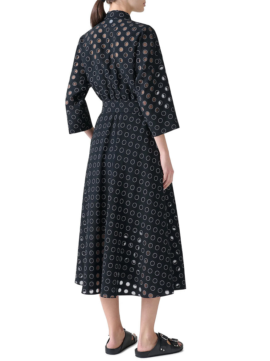 A woman stands facing away, wearing the Akris Punto Midi Polka Dot Organzino Belted Dress in Black/Cream with black sandals and bracelet-length sleeves.
