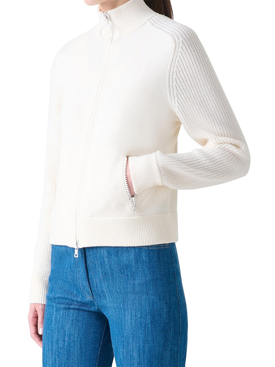 A person wears the Akris Punto Rib-Knit Wool Zip Cardigan in Cream, featuring ribbed sleeves and a stand collar, paired with blue jeans, standing with one hand in a pocket against a white background.