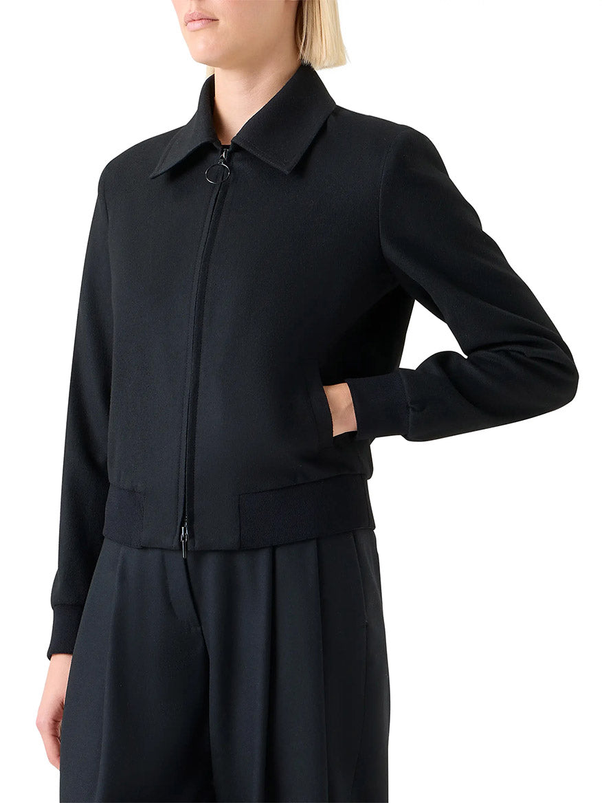 A person wears the Akris Punto Wool/Cashmere Short Bomber Jacket in Black, featuring rib-knit trim, standing with one hand in a pocket against a plain background.