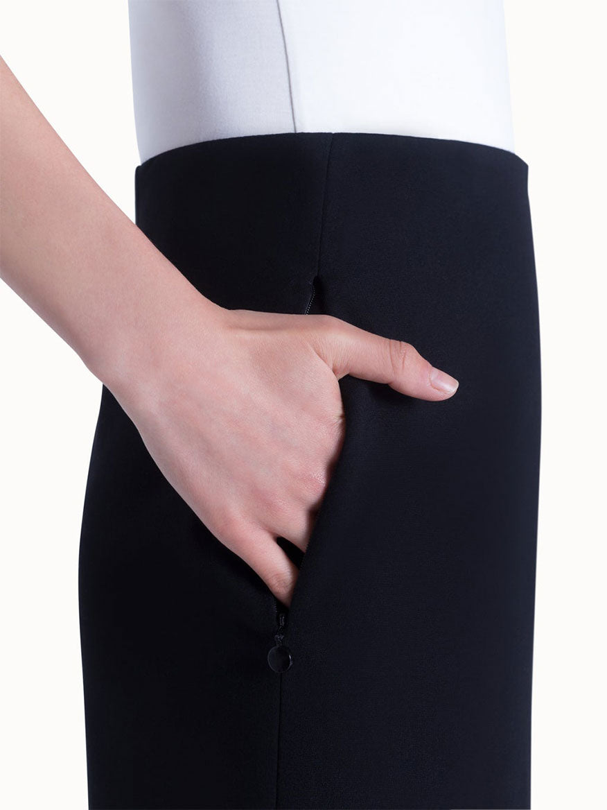 A person places their right hand into the side zipped pocket of the Akris Punto Jersey Stretch Skirt in Black, shown from the waist down against a white background.