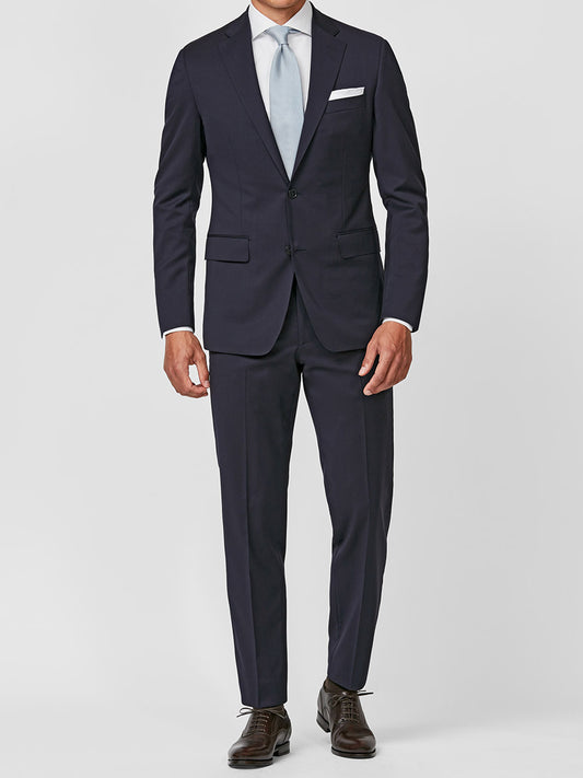 A man wears the Atelier Munro Midnight Blue Twill 4-Season Wool Stretch Suit, paired with a light blue tie, white shirt, white pocket square, and brown dress shoes, standing against a plain light background.