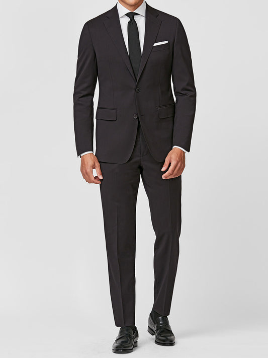 A man wears the Atelier Munro Black Twill 4-Season Wool Stretch Suit with a white dress shirt, black tie, white pocket square, and black dress shoes, standing against a plain white background.