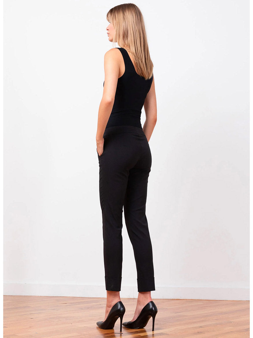 A person with long hair stands in a side profile view, wearing a black sleeveless top, Avenue Montaigne Ben Signature Straight Leg Pants in Black, and high-heeled shoes on a wooden floor against a plain white background.