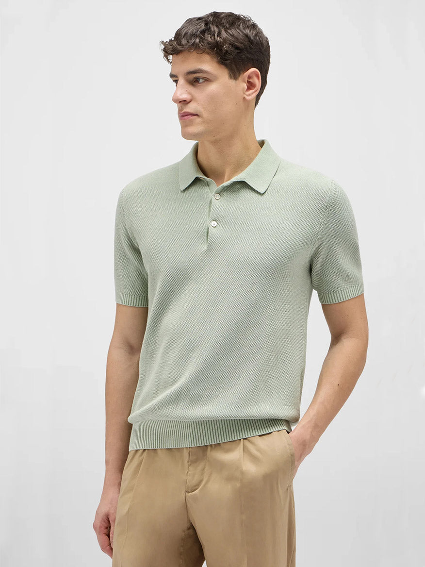 A man wears the Maurizio Baldassari Garment Dyed Cotton Polo Honeycomb Stitch in Aqua Sky with beige pants, standing against a plain light background and looking to the side.