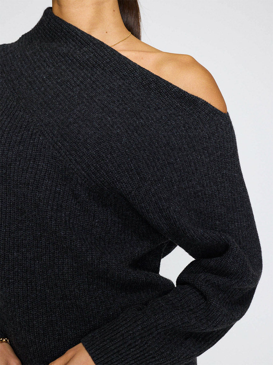 A person wears the Brochu Walker Hart Off-Shoulder Sweater in dark charcoal, made from soft Italian wool cashmere, revealing their left shoulder and part of their arm against a plain background.