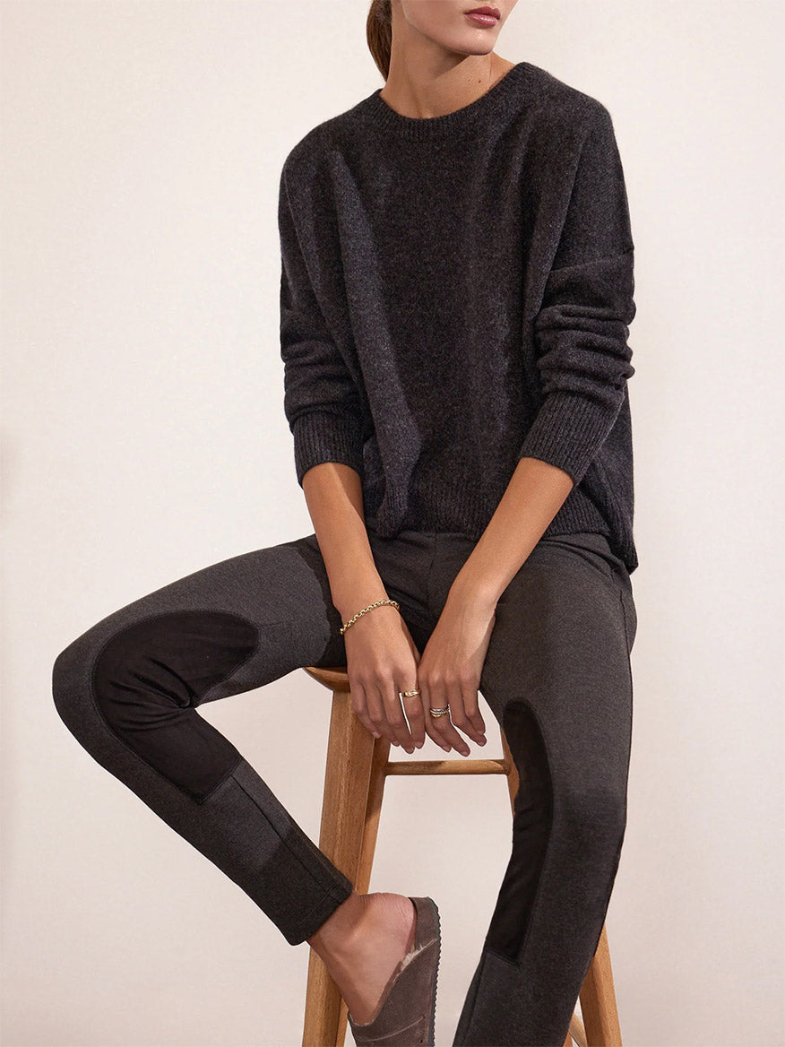 Woman seated on a wooden stool wearing the Brochu Walker Remington Riding Pant in Charcoal Melange and a dark grey sweater featuring vegan leather panels.