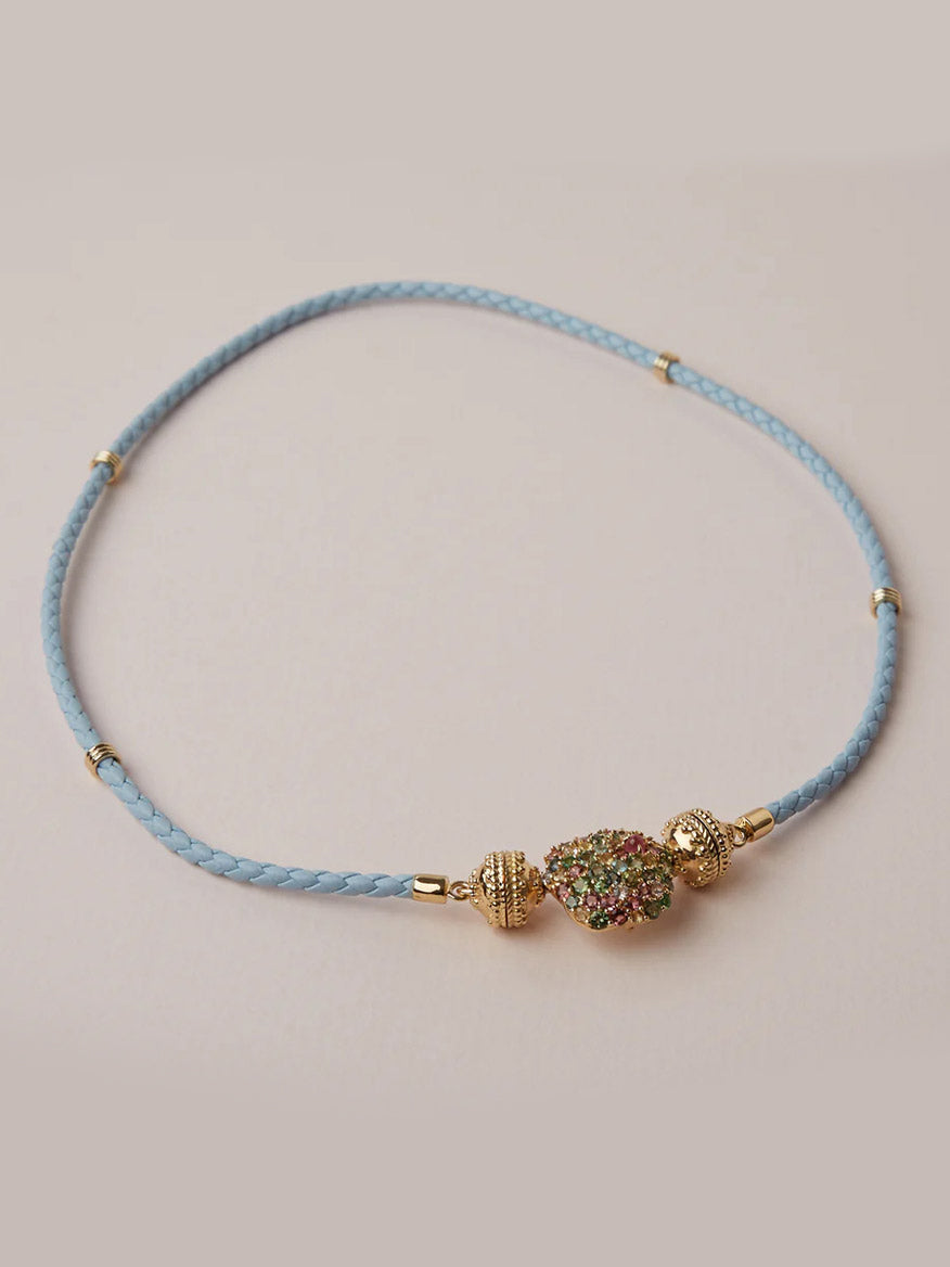 The Clara Williams Aspen Braided Light Blue Leather Mini Necklace features gold-toned beads and a multicolored stone cluster with a ClaraClasp, shown on a plain background.