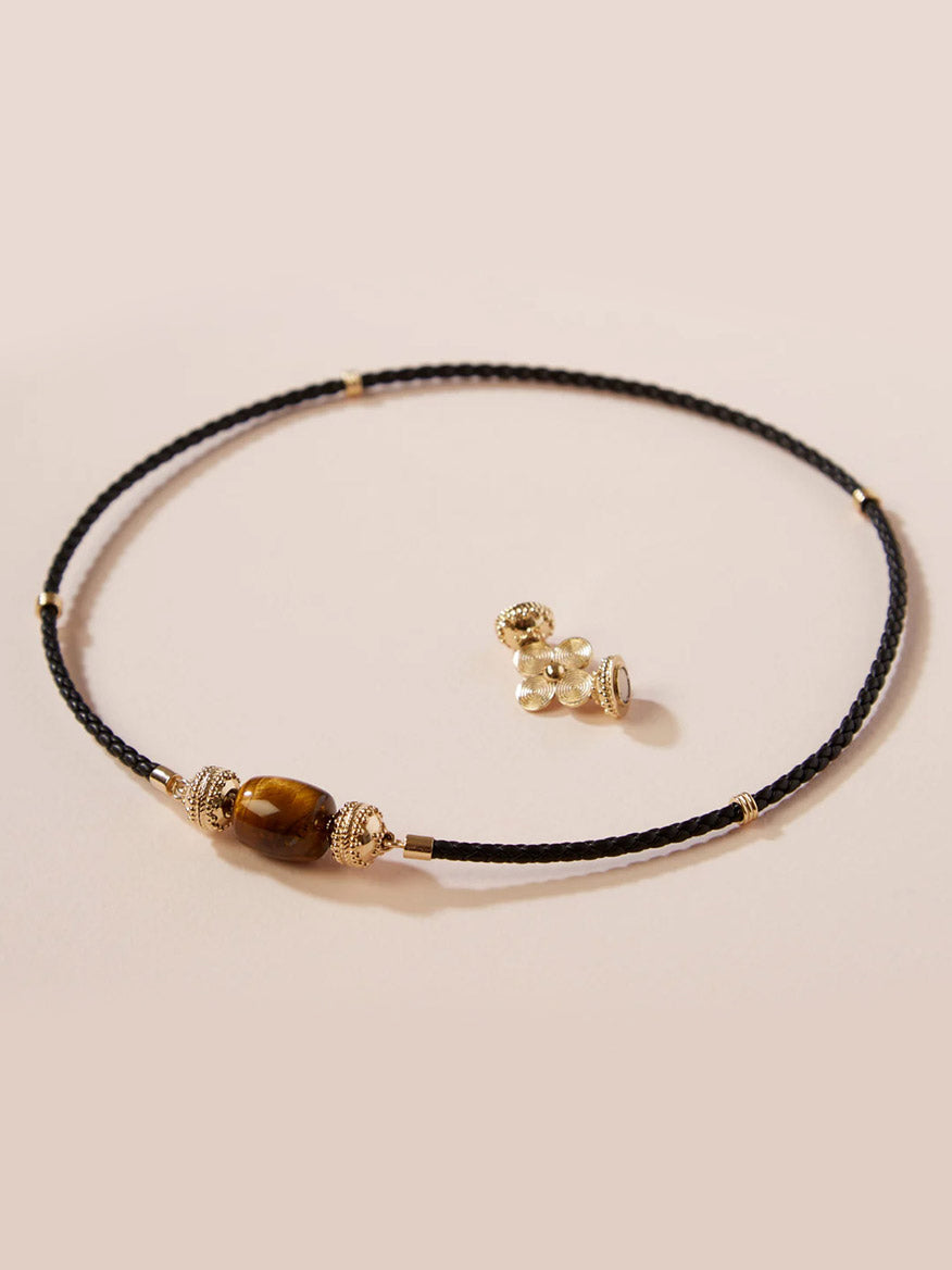 The Clara Williams Aspen Braided Leather Black Mini Necklace features a central brown gemstone bead, gold vermeil accents, and a sterling silver clasp, shown beside a small gold-toned floral stud earring.