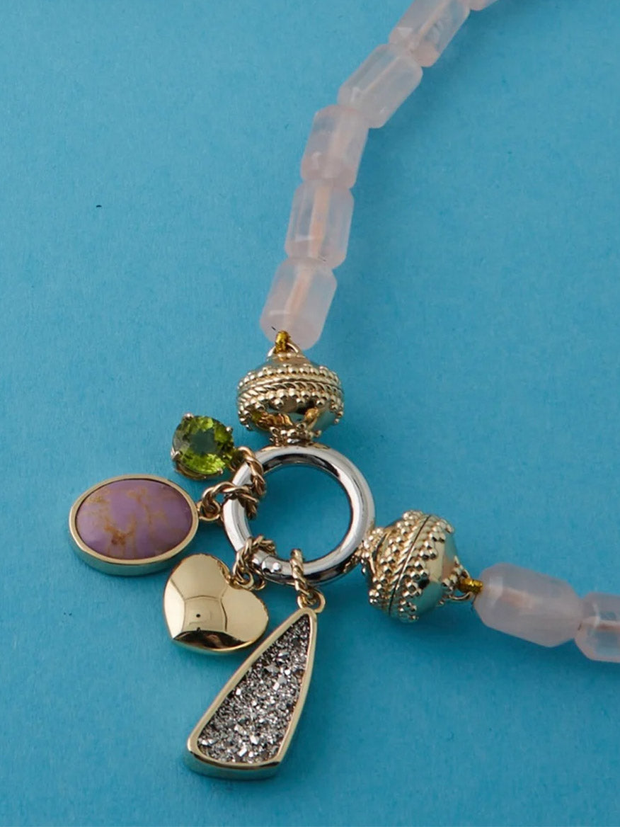 The Clara Williams ClaraCharm™ Rhodium Ring Mini Centerpiece is a beaded mini necklace with a central ring and five interchangeable charms—pink stone, green gem, textured silver, heart, and round bead—set on a light blue background.