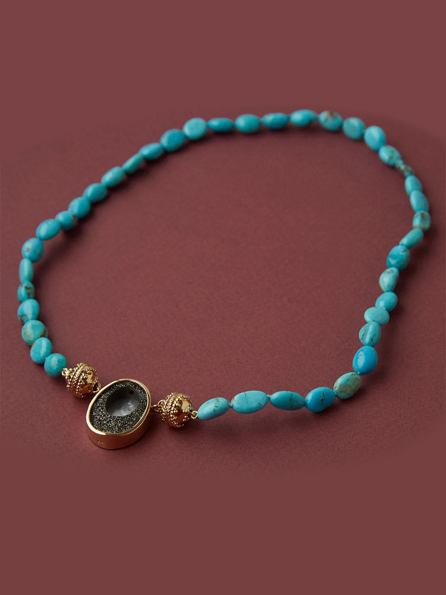 The Clara Williams 14K Black Druzy Oval Cut-Out Mini Centerpiece features a turquoise beaded necklace with yellow gold accents, shown on a maroon background.