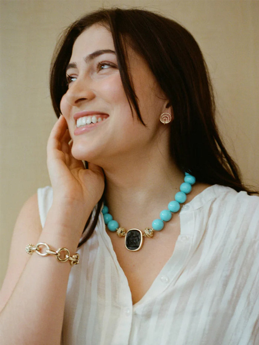 Woman with long dark hair smiles in a sleeveless white top, Clara Williams turquoise beaded necklace with black pendant, Gold Coast Bracelet in 14K yellow gold, and circular gold earrings against a neutral background.